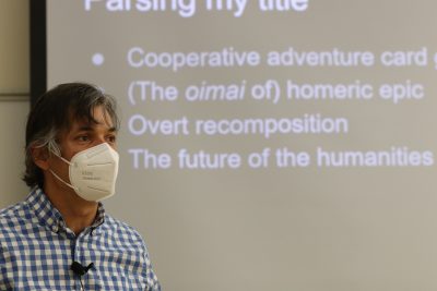 Roger Travis (wearing a white and blue checkered collared shirt and white mask) presents about recomposition; slides projected behind him say "The future of the humanities"